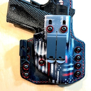 Peut inclure: Un étui d'arme à feu noir et gris avec un motif de drapeau américain rouge et blanc. L'étui a un clip noir et des accents rouges. La couture est rouge.