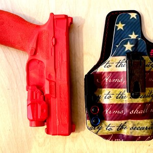 Pu&ograve; includere: Una pistola da addestramento rosso vivo e una fondina fantasia. La fondina presenta un disegno della bandiera americana con stelle, strisce e testo. La pistola &egrave; di colore rosso pieno.
