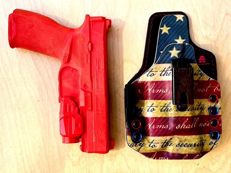 Pu&ograve; includere: Una pistola da addestramento rosso vivo e una fondina fantasia. La fondina presenta un disegno della bandiera americana con stelle, strisce e testo. La pistola &egrave; di colore rosso pieno.