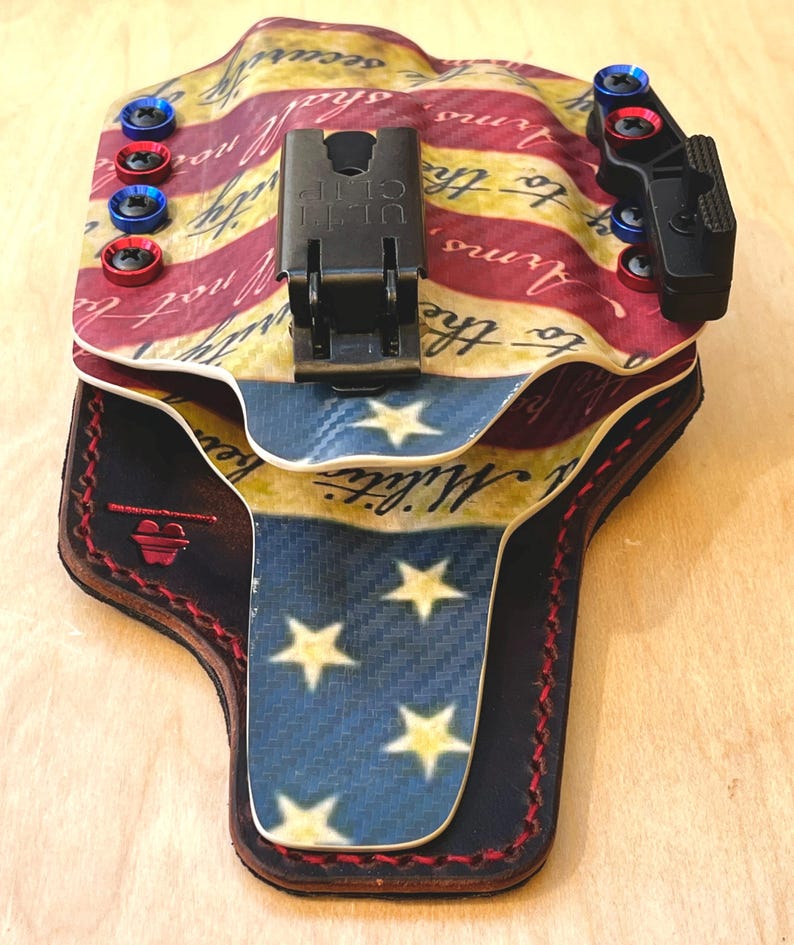 Pu&ograve; includere: Una fondina personalizzata con un design a bandiera americana. La fondina ha una combinazione di colori rosso, bianco e blu con testo e stelle. Include una clip nera e viti rosse e blu.