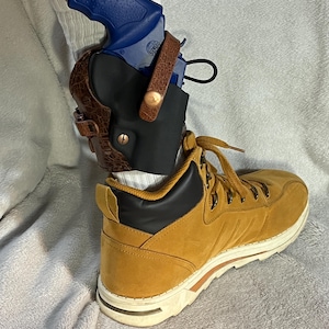 Puede incluir: Una pistola azul en una funda de cuero marrón, sujeta a la pierna. La funda está sujeta a una bota marrón con cuello negro y suela blanca. La bota está atada con cordones marrones.