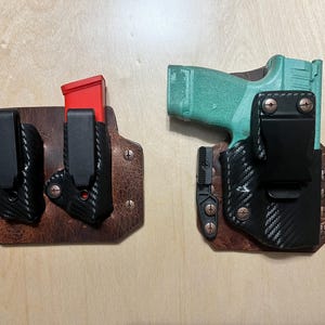 Op de afbeelding: Twee bruine leren holster met zwarte en rode accenten. Een holster bevat een magazijn en de andere een pistool. Het pistool is turquoise en zwart.