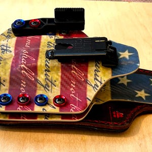 Pu&ograve; includere: Una fondina personalizzata con un design patriottico nei colori rosso, bianco e blu. La fondina presenta un motivo a tema bandiera con stelle e strisce, ed &egrave; visibile il testo "the security of". Include clip nere e viti rosse e blu.