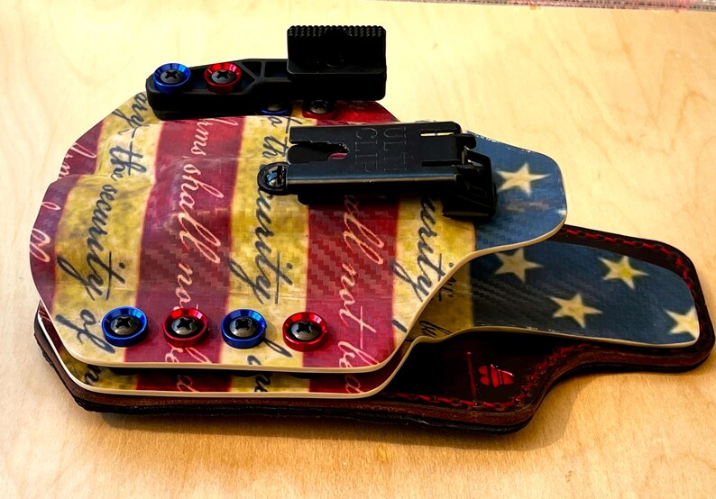 Pu&ograve; includere: Una fondina personalizzata con un design patriottico nei colori rosso, bianco e blu. La fondina presenta un motivo a tema bandiera con stelle e strisce, ed &egrave; visibile il testo "the security of". Include clip nere e viti rosse e blu.