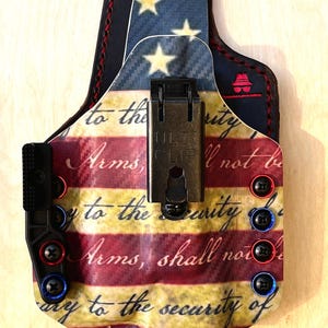 Pu&ograve; includere: Una fondina personalizzata con un design a bandiera americana. La fondina ha una combinazione di colori rosso, bianco e blu con il testo "Arms, shall not be" e "to the security of". Include una clip nera e viti rosse e blu.