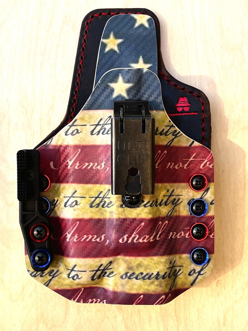 Pu&ograve; includere: Una fondina personalizzata con un design a bandiera americana. La fondina ha una combinazione di colori rosso, bianco e blu con il testo "Arms, shall not be" e "to the security of". Include una clip nera e viti rosse e blu.