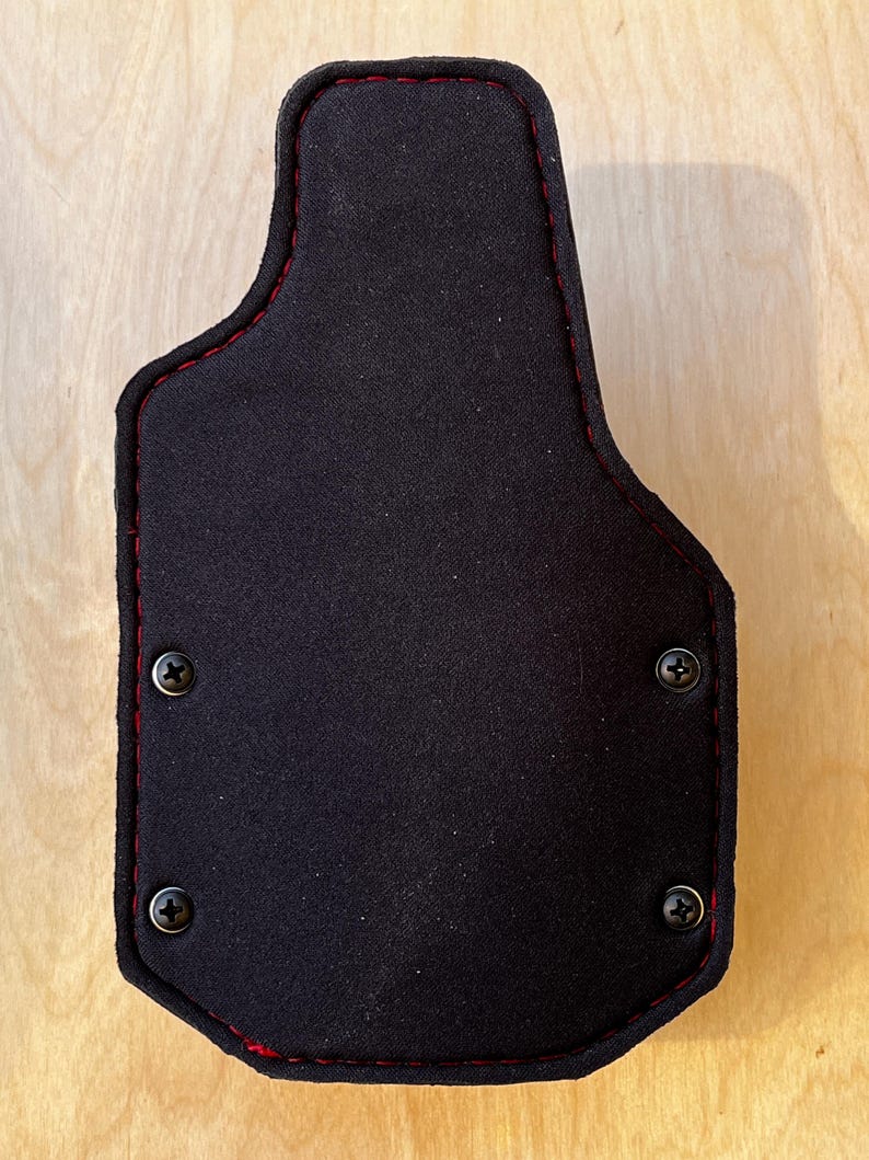 Pu&ograve; includere: Una fondina nera con cuciture rosse sui bordi. La fondina ha una forma sagomata ed &egrave; fissata con quattro viti nere. Lo sfondo &egrave; una venatura del legno chiaro.