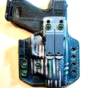 Peut inclure: Un étui noir et bleu avec une fine bande verte et un motif d'étoiles et de rayures blanches. L'étui a des vis vertes et un clip noir. L'étui est conçu pour contenir une arme de poing.