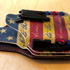 Pu&ograve; includere: Una fondina multistrato con un design a bandiera americana. La fondina presenta una sezione blu con stelle dorate e una sezione a strisce rosse, bianche e gialle con testo. Ha hardware nero e accenti rossi e blu.