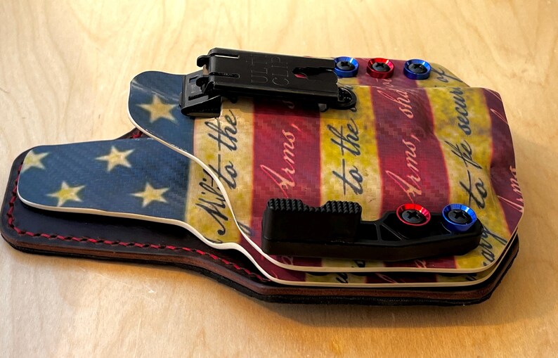 Pu&ograve; includere: Una fondina multistrato con un design a bandiera americana. La fondina presenta una sezione blu con stelle dorate e una sezione a strisce rosse, bianche e gialle con testo. Ha hardware nero e accenti rossi e blu.