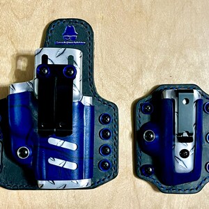 May include: A holster and a magazine carrier set featuring a blue and silver diamond plate pattern. Both items are mounted on black leather backings with black metal belt clips and blue hardware accents. A blue logo is visible on the holster.