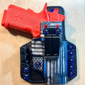 Peut inclure: Un pistolet rouge est posé dans un étui noir avec un motif de drapeau américain bleu et blanc. L'étui comporte des vis bleues et un clip noir. Le pistolet a une poignée texturée et un motif d'étoile.