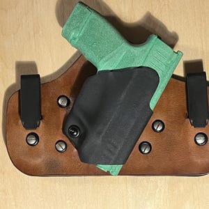 Op de afbeelding: Een bruin leren holster met een zwart interieur en zwarte riemclips. Een groen pistool is gedeeltelijk in het holster gestoken. Het holster is vastgezet met zilveren klinknagels.