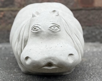 Puede incluir: Una escultura de hipopótamo de hormigón gris claro. El hipopótamo tiene ojos, fosas nasales y una boca ancha detallados. La escultura es una forma sólida y redondeada, adecuada para la decoración exterior o interior. El hipopótamo es una adición caprichosa a cualquier jardín o hogar.