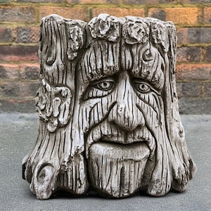 Op de afbeelding: Een grijze betonnen plantenbak in de vorm van een boomstronk met een gezicht erin gegraveerd. Het gezicht heeft een lange baard, een brede mond en twee ogen. De plantenbak is versierd met kleine, gegraveerde bladeren.