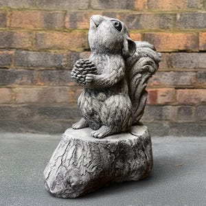 Puede incluir: Estatua de ardilla gris sosteniendo una piña, posada en un tocón de árbol. La ardilla mira hacia arriba. La estatua está hecha de un material similar a la piedra y es adecuada para la decoración exterior.