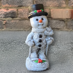 May include: A snowman figurine with a black top hat adorned with holly and berries. The snowman has a carrot nose, black button eyes, and a scarf. It holds a cane and stands on a base decorated with holly.
