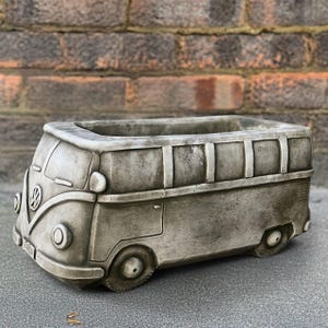 May include: A gray concrete planter shaped like a vintage Volkswagen bus. The planter has a rectangular opening for plants, with details including headlights, wheels, and the VW logo. The planter is designed to sit on a flat surface.