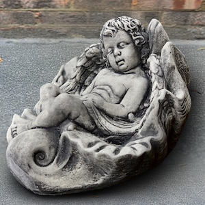 Puede incluir: Escultura de piedra gris de un querubín durmiente, acunado en una forma de concha. El querubín tiene alas detalladas y cabello rizado. Escultura decorativa para jardín.