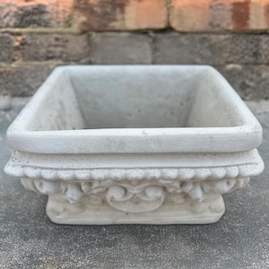 May include: A white square planter with an ornate design around the top edge. The planter is made of concrete and has a smooth finish.