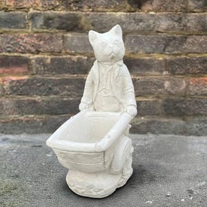 May include: A light-colored cat statue pushing a wheelbarrow planter. The cat is wearing a jacket and vest. The wheelbarrow has a wooden design. The statue is made of a light-colored material and is suitable for outdoor use.