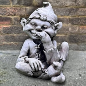May include: A gray garden gnome statue with a mischievous expression. The gnome is seated, wearing a pointed hat and tunic, with one hand raised to its mouth. The statue has detailed features and a weathered appearance.