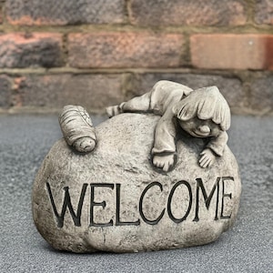 Puede incluir: Adorno de jardín de piedra gris con una figura de niño descansando sobre una roca. La palabra "WELCOME" está tallada en la roca. El adorno tiene un aspecto envejecido, lo que sugiere su uso en exteriores.