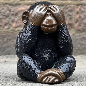 May include: A black ceramic monkey figurine with brown hands covering its eyes. The monkey is sitting on a gray surface.