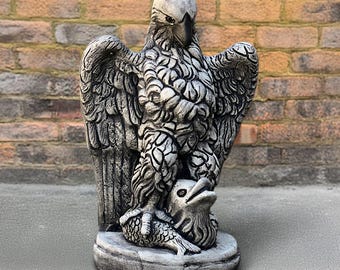 May include: A detailed, gray concrete eagle statue with outstretched wings, perched atop a fish. The eagle's feathers are intricately carved, and the statue has a weathered appearance, suitable for outdoor decor.