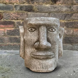May include: A weathered, stone-effect planter sculpted in the form of a human face. The grey sculpture has distinct facial features, including eyes, a nose, and a mouth. The planter is designed for plants and adds a unique decorative element.