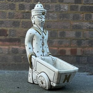 May include: White ceramic figurine of a person wearing a hat and robe, holding a white wheelbarrow with a gold handle. The wheelbarrow has a gold letter 'H' on the side.