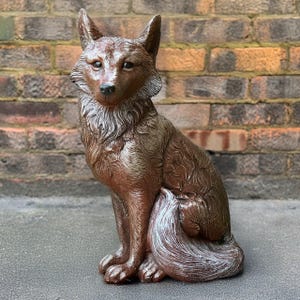May include: A brown and white ceramic wolf figurine sitting on a brick surface. The wolf is sitting with its tail curled around its legs. The figurine is detailed with fur and has a realistic look.