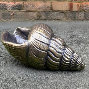 Könnte beinhalten: Eine bronzefarbene Muschelbildhauerei mit detaillierten Merkmalen. Die Muschel hat eine strukturierte Oberfläche mit ausgeprägten Rippen und Kurven. Sie liegt auf einer grauen Oberfläche. Die Öffnung der Muschel ist sichtbar und erinnert an eine natürliche Muschel.