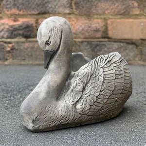 Puede incluir: Una maceta de cisne de hormigón gris con texturas detalladas de plumas y alas. El cisne tiene un cuello curvado y una cabeza mirando hacia abajo. La maceta está diseñada para contener plantas y añade un toque decorativo a jardines o patios.