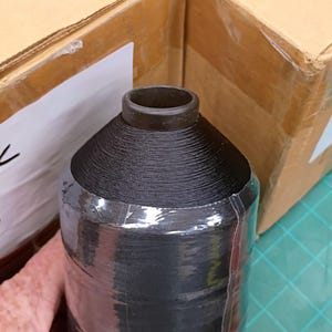May include: A large spool of black thread featuring a dark brown plastic core at the top. The thread is tightly wound in a cylindrical shape and partially wrapped in a clear, thin plastic film that covers the lower portion of the spool.