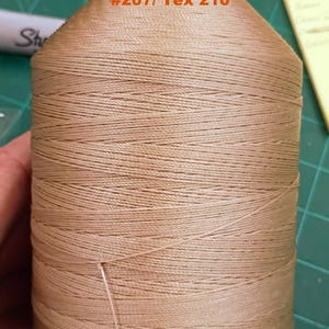 May include: A large spool of beige thread with a black top and bottom. The text printed on the upper portion of the thread reads Beige #207/ Tex 210. The thread is wound tightly around the cylindrical spool, showing a consistent light brown color.