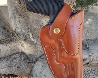 Könnte beinhalten: Eine braune Lederholster mit einer schwarzen Handfeuerwaffe. Die Holster hat eine glänzende Oberfläche, dekorative Nähte und einen Messingknopfverschluss. Der Text "Big John's Holsters And Sheaths" ist unten sichtbar.