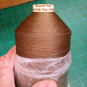 May include: A large spool of desert tan colored thread. The white label on the top of the spool features the text Desert Tan #138/ Tex 135. The lower portion of the spool is wrapped in a clear plastic film, showing the tightly wound brown thread.