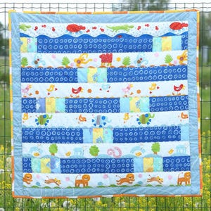 Op de afbeelding: Een blauwe en witte babydeken met een patchwork-ontwerp met verschillende dieren, waaronder leeuwen, olifanten, vogels en vissen. De deken heeft een lichtblauwe achtergrond met witte cirkels en een rand van oranje en witte strepen.