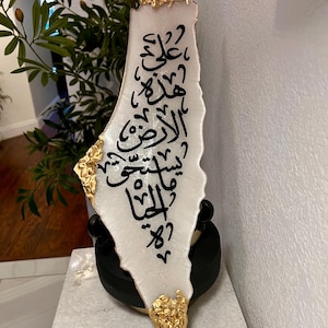 May include: A white ceramic outline of the country of Palestine with gold accents. The outline is decorated with Arabic calligraphy in black.