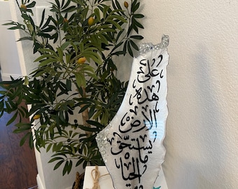 Könnte beinhalten: Eine silberne Umrandung des Landes Palästina mit arabischer Kalligraphie in Schwarz. Die Umrandung ist mit silbernem Glitzer verziert. Die Umrandung steht auf einer weißen Holzkiste.