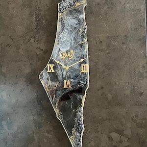 Op de afbeelding: Een decoratieve wandklok in de vorm van een kaart, met een zwart-wit marmerpatroon en gouden accenten. De klok heeft gouden Romeinse cijfers en wijzers, met Arabisch schrift in het midden. De randen zijn getextureerd met zwart en goud.