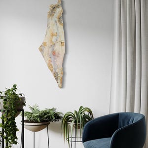 May include: A wall-mounted art piece in the shape of a country, featuring a mix of beige, gold, and pink tones. The artwork has a textured surface and includes what appears to be Arabic script. The piece is hanging on a white wall.