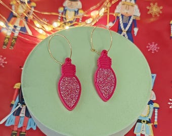 May include: A pair of festive earrings featuring a pink Christmas light bulb design with glitter accents. The earrings hang from gold-colored hoops. The earrings are displayed on a light green surface against a red background with nutcracker illustrations.