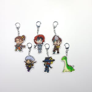 May include: Set of seven colourful cartoon character keychains. The keychains feature a variety of characters, including a dinosaur, a person wearing a hat, and a person with a gun. The keychains are made of clear acrylic and have silver metal rings.