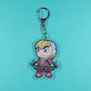 Puede incluir: Un llavero con un personaje de dibujos animados con cabello rubio, que lleva una chaqueta rosa y morada y un casco blanco. El personaje est&aacute; extendiendo las manos, con el texto "Let's Get Weird" en el llavero.