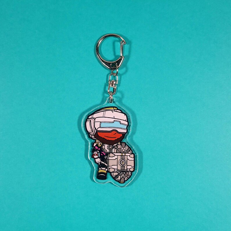 Op de afbeelding: Een wit en grijs cartoonfiguur met een rood gezicht en een witte helm die een grijs en wit rond object vasthoudt. Het figuur is bevestigd aan een zilveren sleutelhanger.