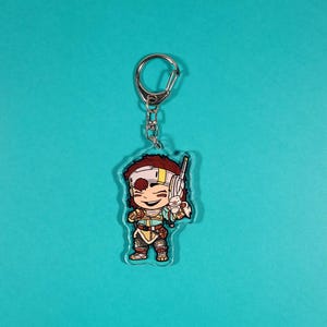 Op de afbeelding: Een transparante acryl sleutelhanger met een cartoonfiguur met bruin haar, een wit hoofdband en een geel shirt. Het personage houdt een klein wit dier in de ene hand en een bruine stok in de andere. De sleutelhanger is bevestigd aan een zilveren metalen ring.