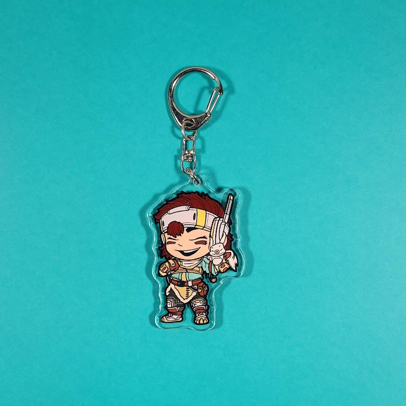 Op de afbeelding: Een transparante acryl sleutelhanger met een cartoonfiguur met bruin haar, een wit hoofdband en een geel shirt. Het personage houdt een klein wit dier in de ene hand en een bruine stok in de andere. De sleutelhanger is bevestigd aan een zilveren metalen ring.