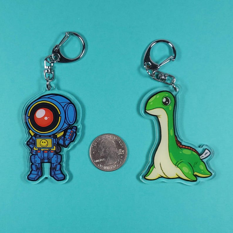 May include: Two acrylic keychains, one featuring a blue robot with a red eye and a yellow smiley face, and the other featuring a green cartoon dinosaur with a long neck.